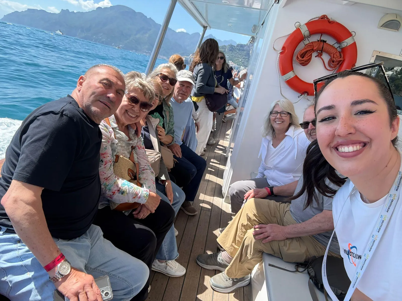 Passengers seated on a boat deck beside a life ring, with mountains across the water