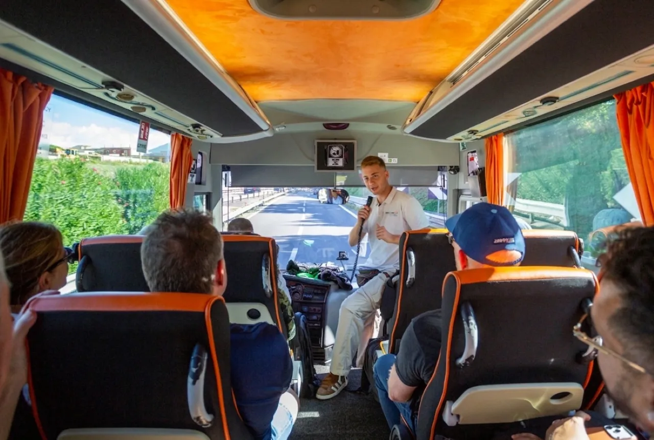 Guide with a microphone at the front of a coach with passengers in orange-trimmed seats