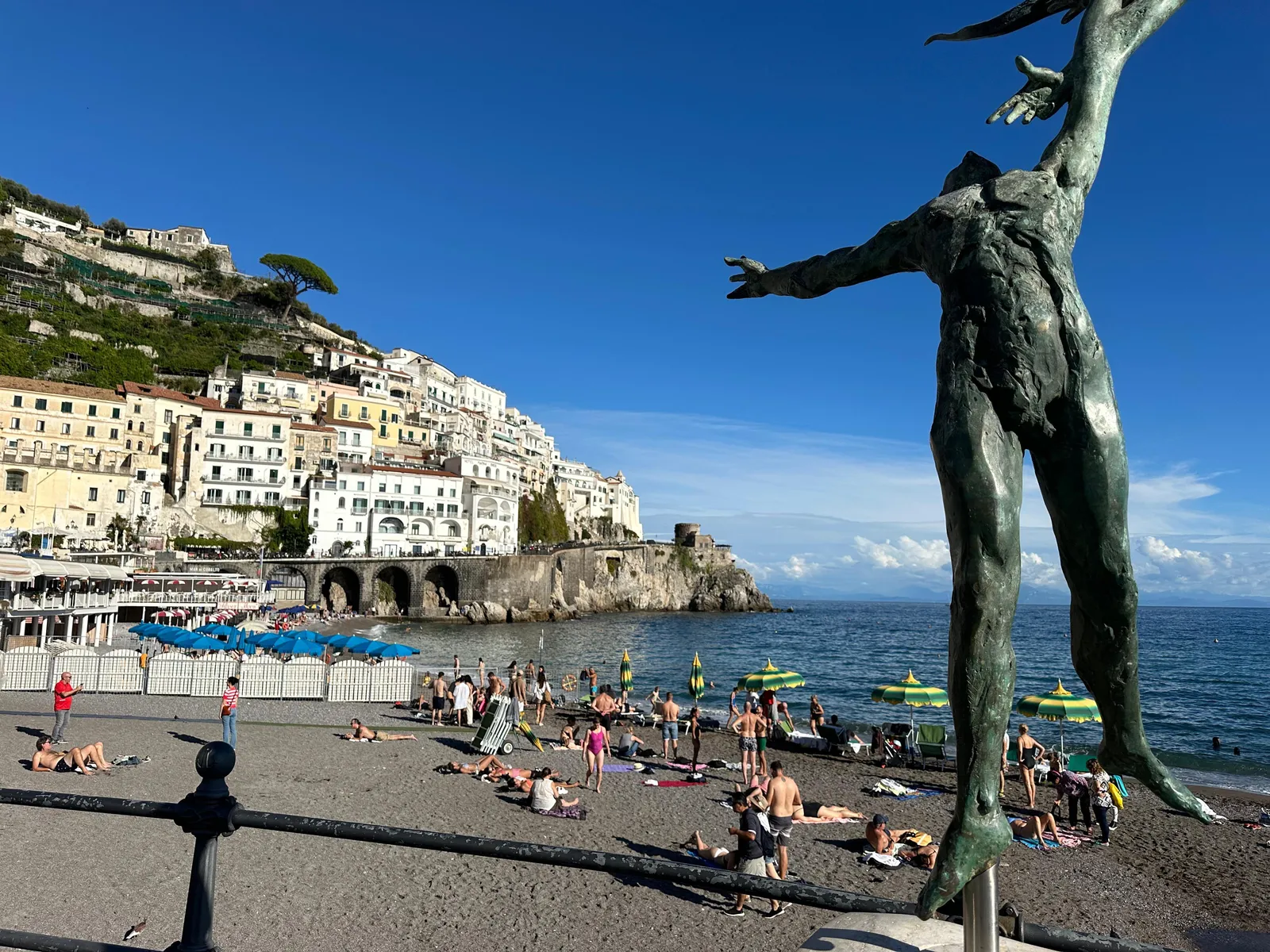 Bronze figure on a seafront promenade above a crowded pebble beach and cliffside town