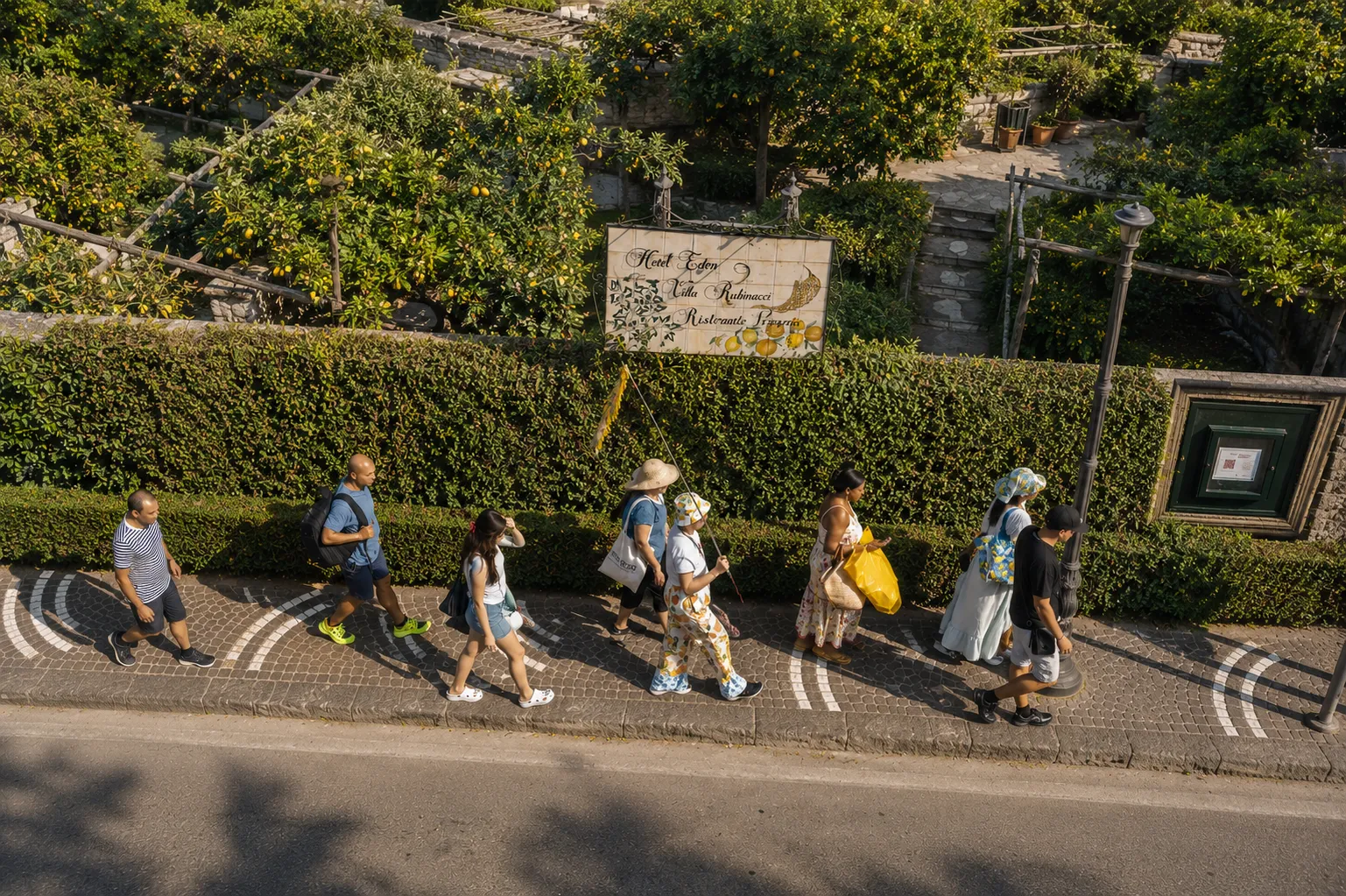 Group of walkers on a paved street beside a hedge and green hillside