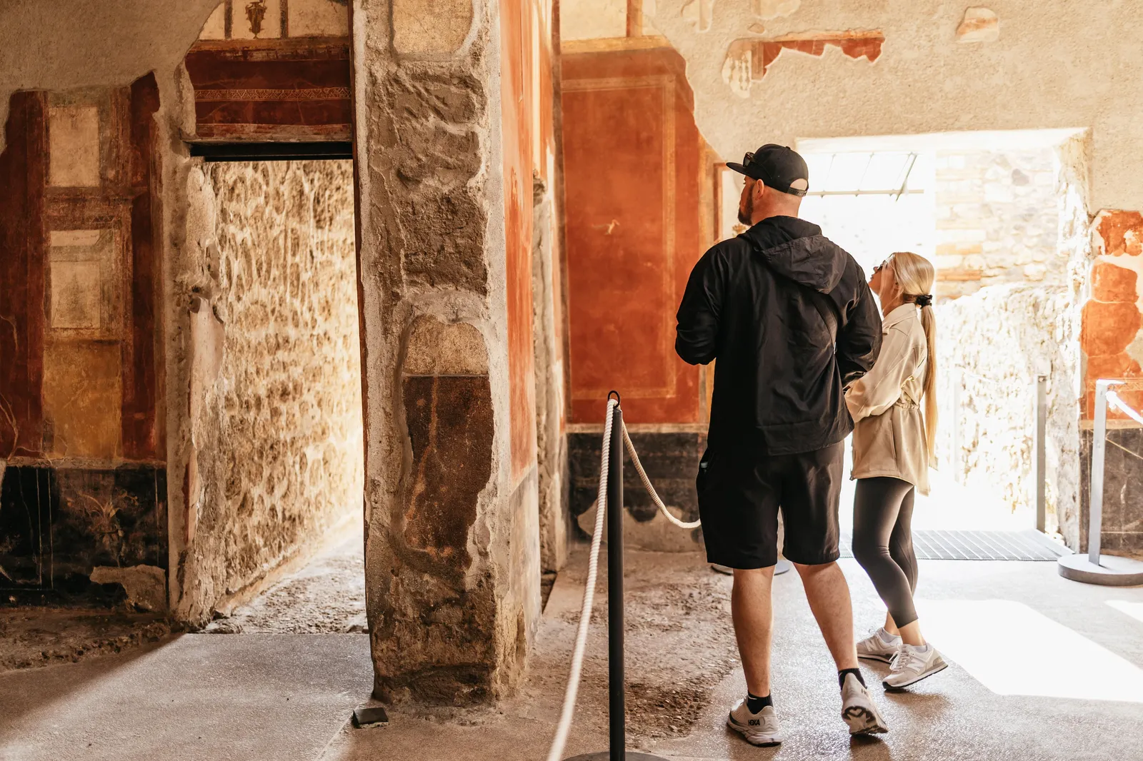 Two visitors walking through a Pompeii house with faded red painted walls