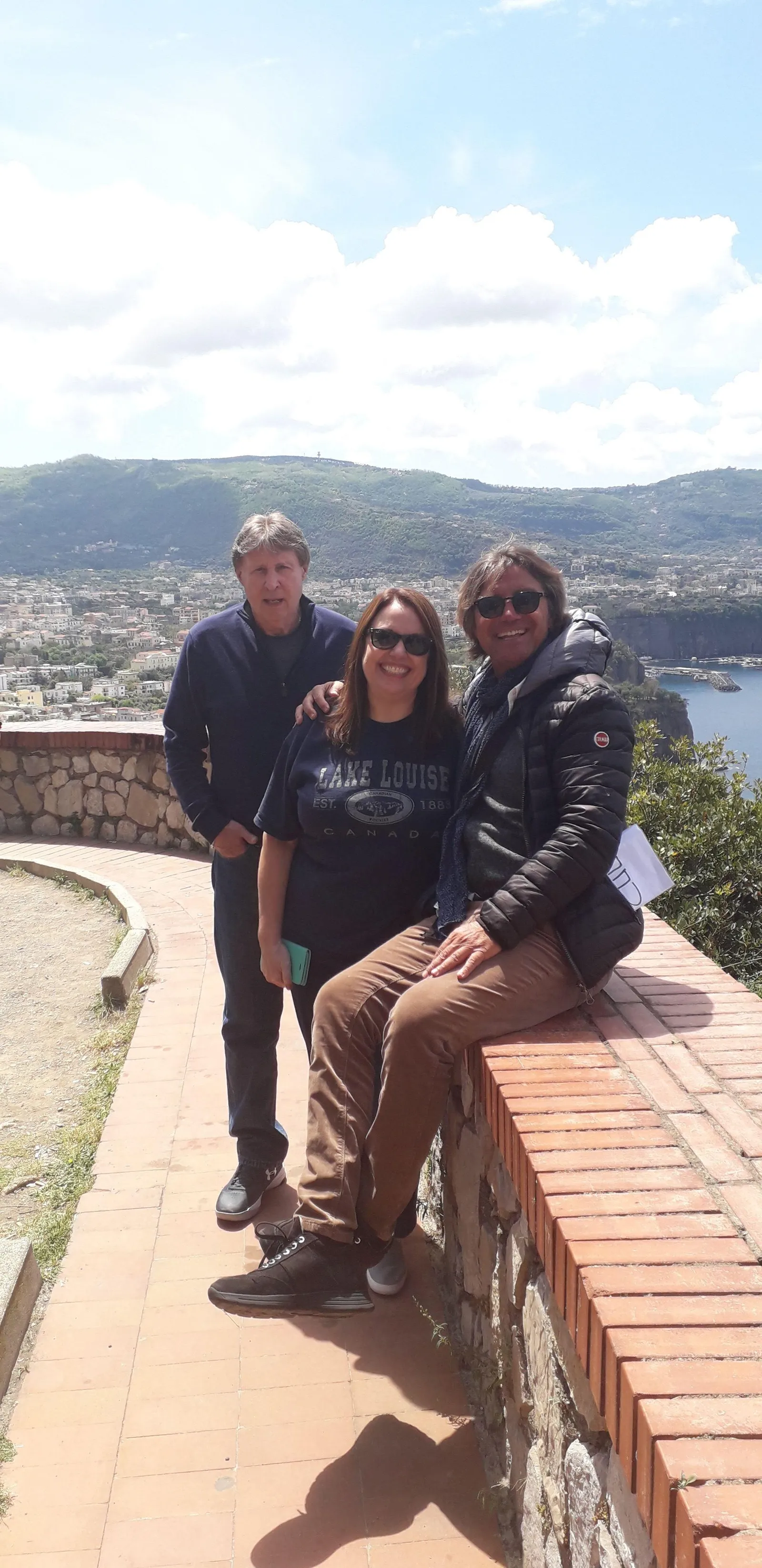 Three people sitting on a stone wall at a viewpoint overlooking a bay