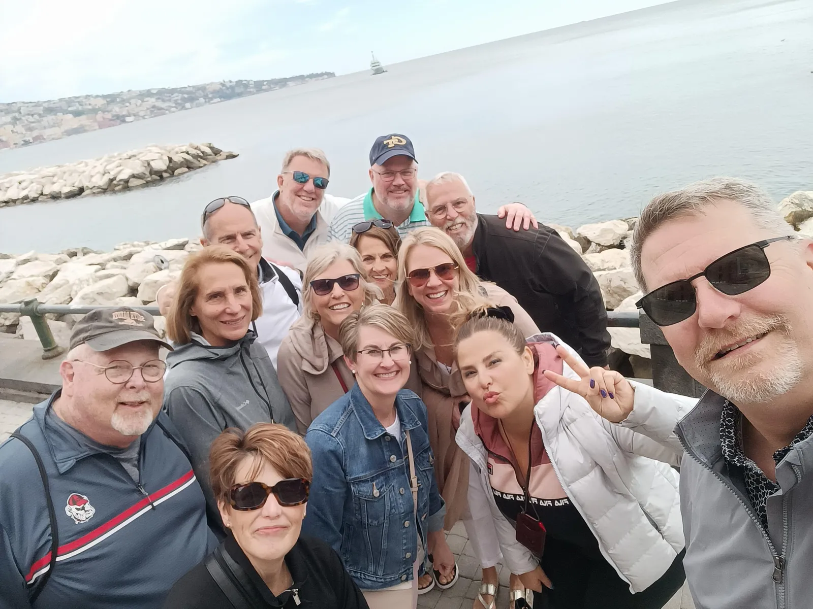 Group selfie on a seafront breakwater with a bay and distant coast behind