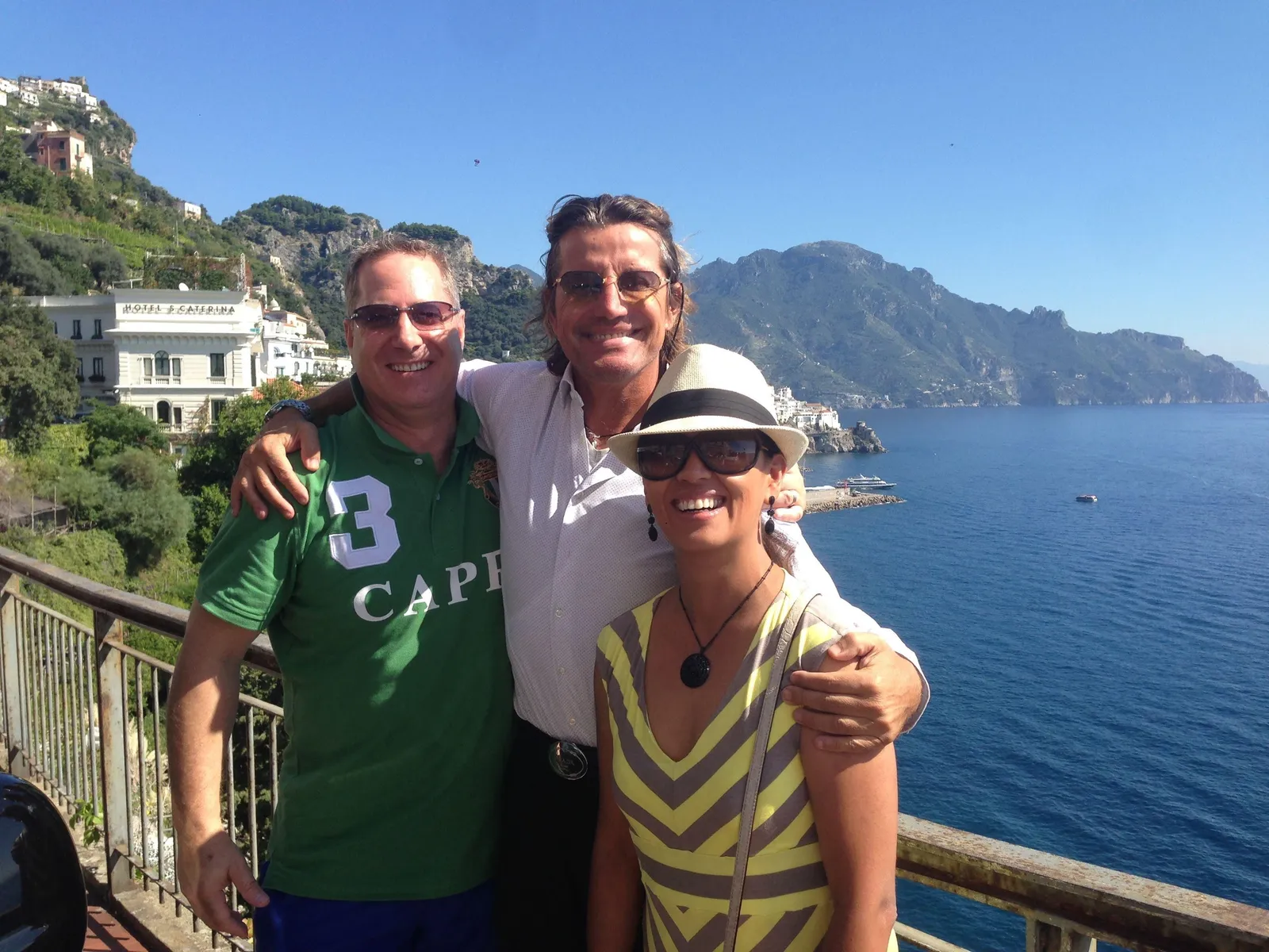 Three people with arms around each other at a railing above the sea and coast