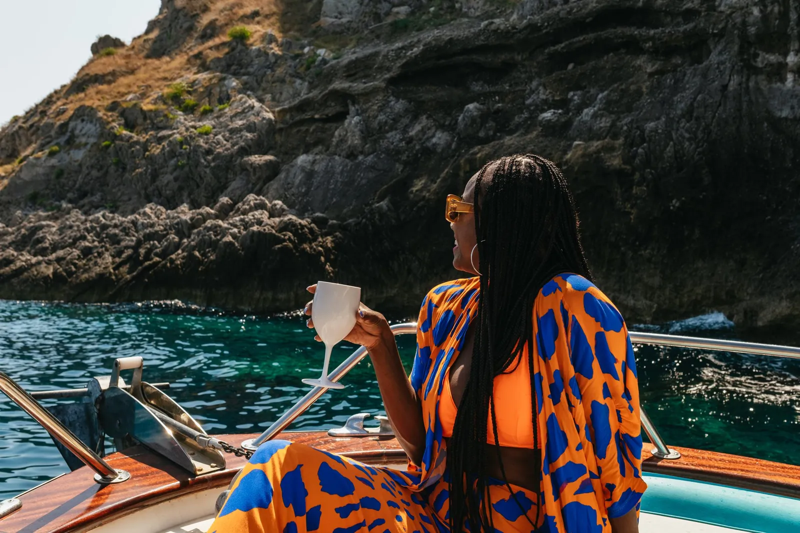 Woman holding a cup on the deck of a wooden boat beside a dark rock cliff