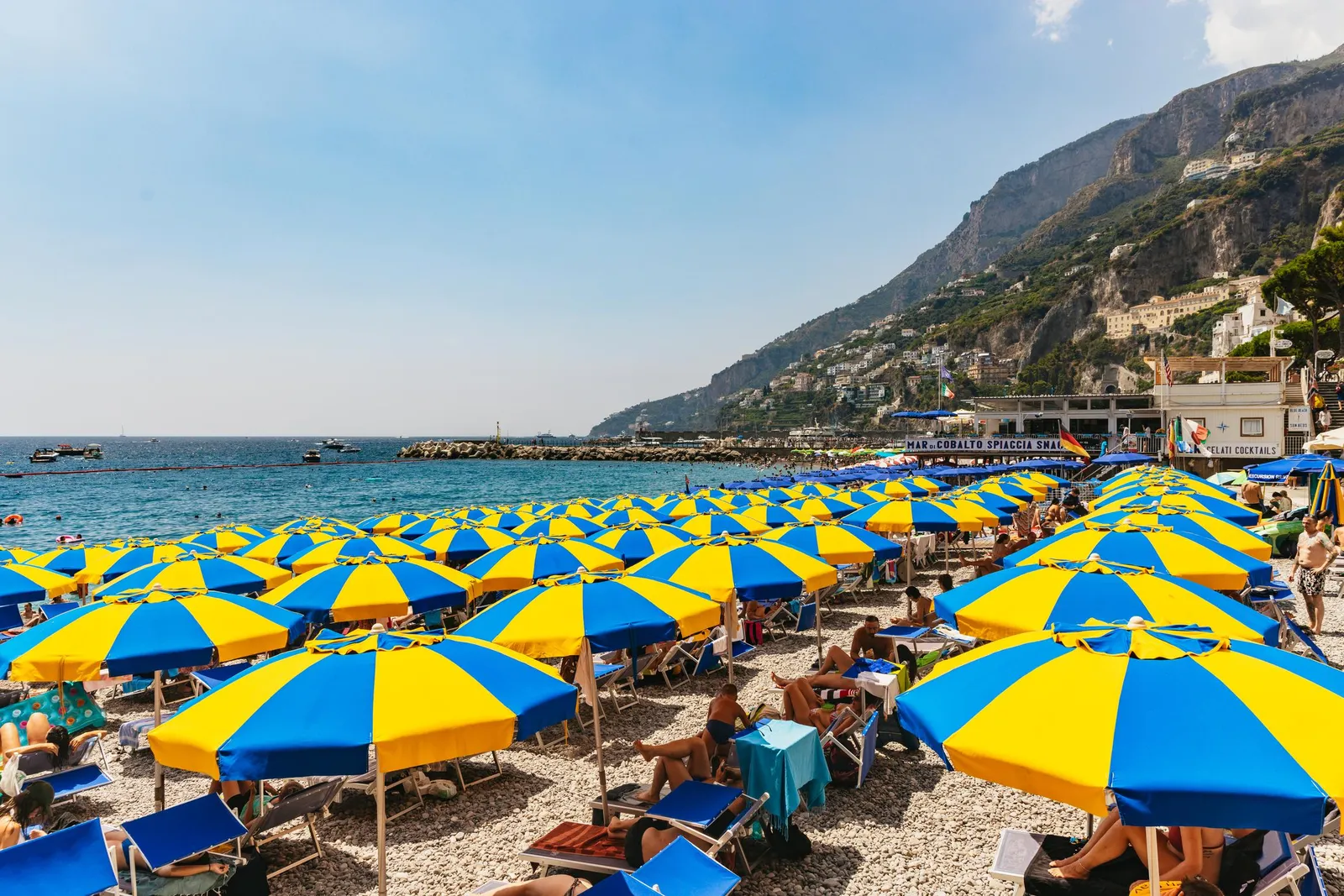 Rows of blue and yellow beach umbrellas on a crowded beach below steep mountains