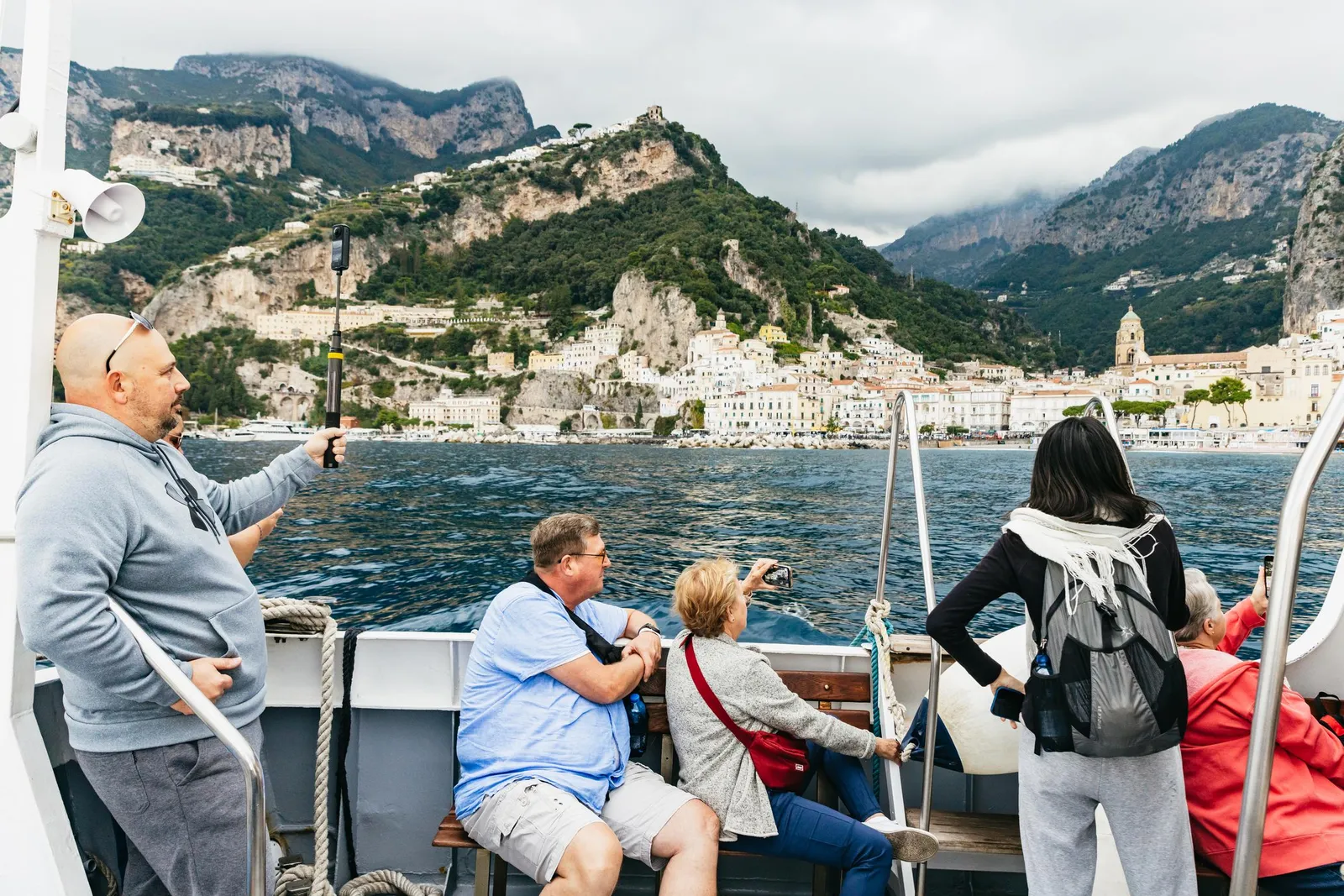 Passengers on a boat deck looking toward a cliffside town and mountains