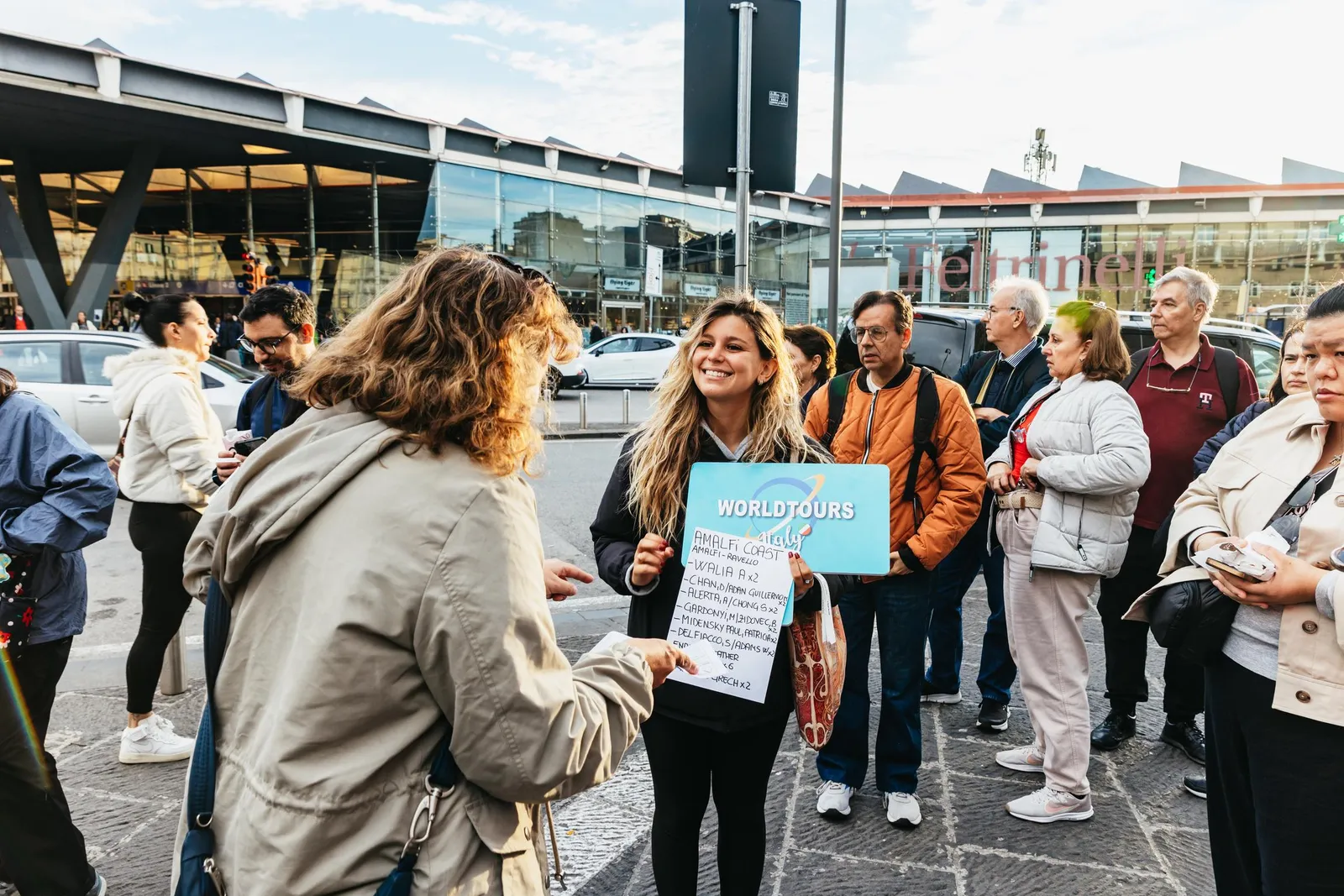 Tour guide holding a blue sign and addressing a group outside a station building