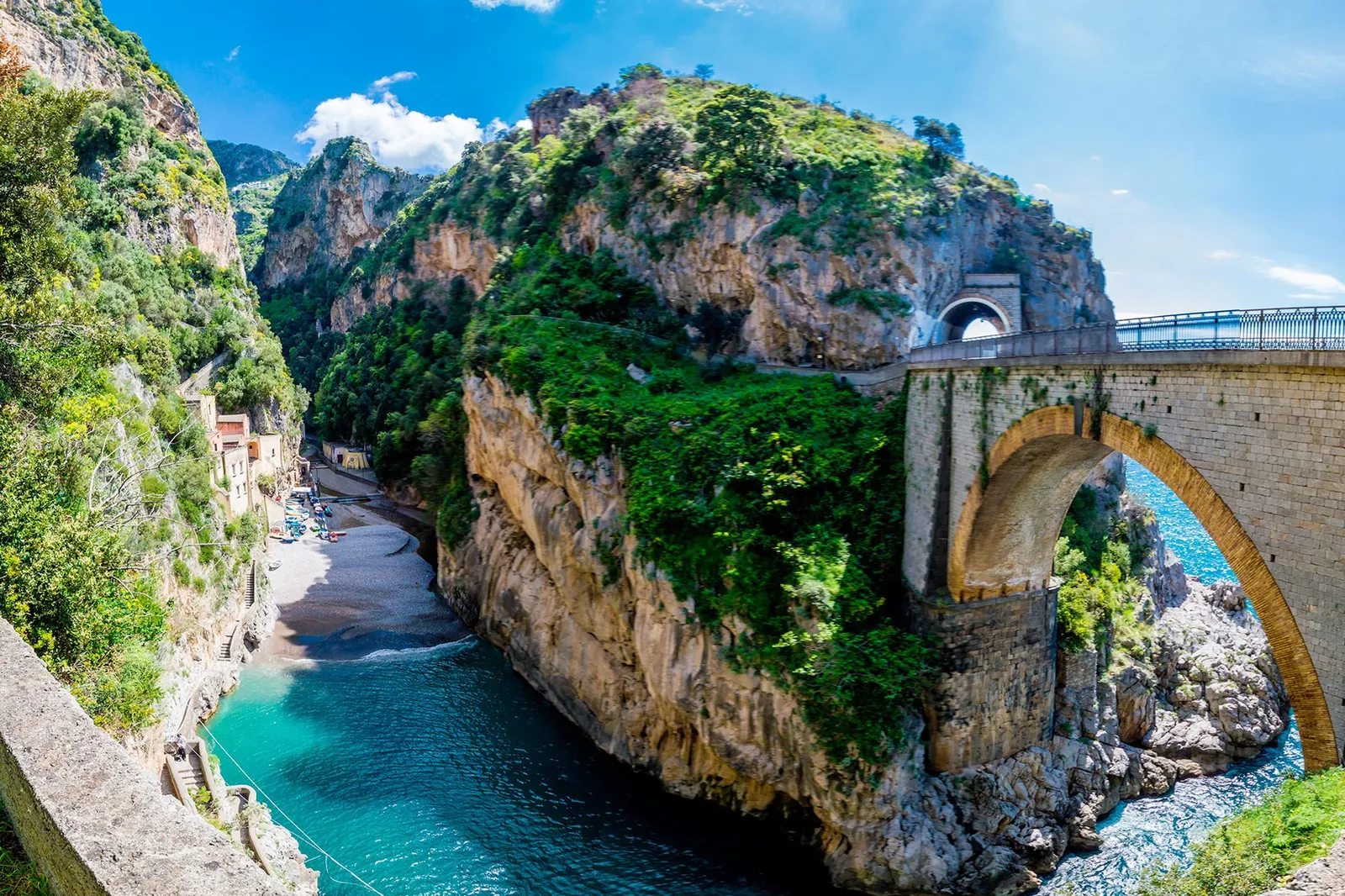 Stone arched bridge over a narrow rocky inlet with a small beach and green cliffs