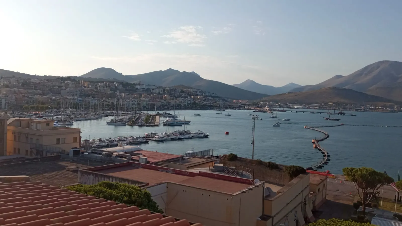 Harbour with moored boats and a breakwater below distant mountains, seen from rooftops