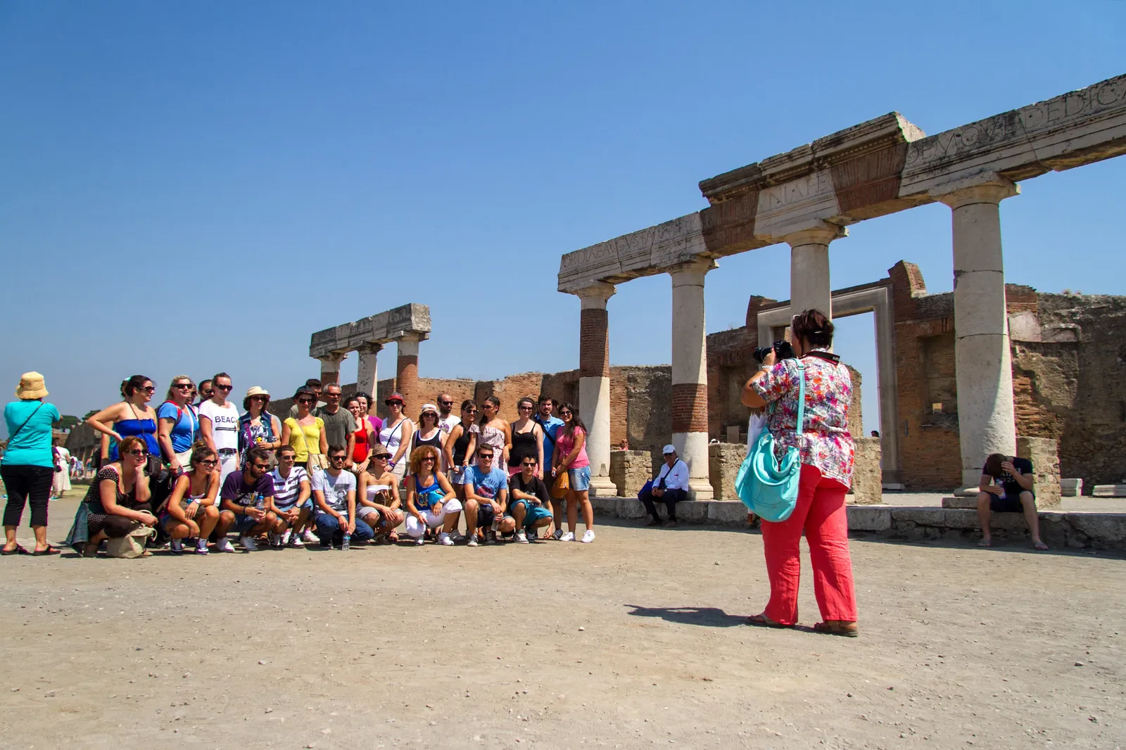 Large group seated for a photo among columns at Pompeii's forum, a woman taking the picture