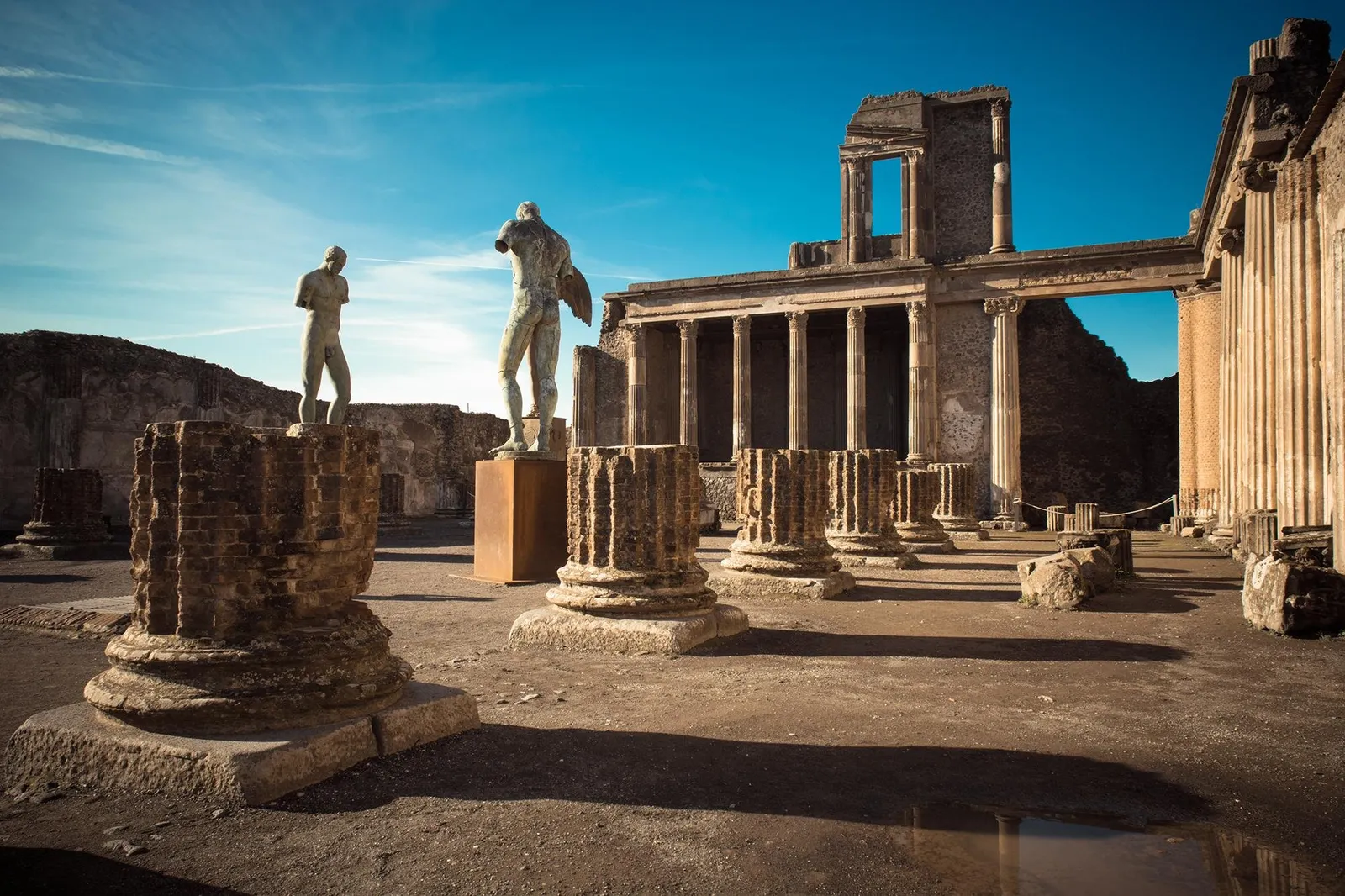 Statues and column bases in a ruined Pompeii building under a blue sky
