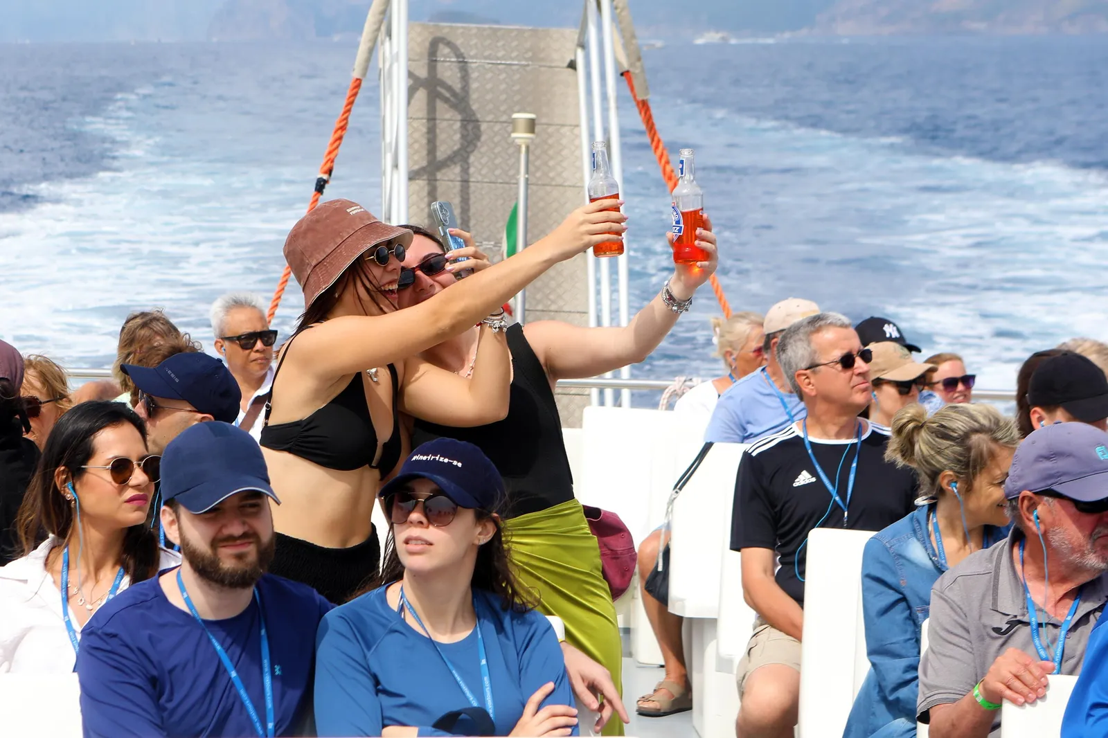 Passengers seated on a boat deck, some holding drinks, with white wake behind