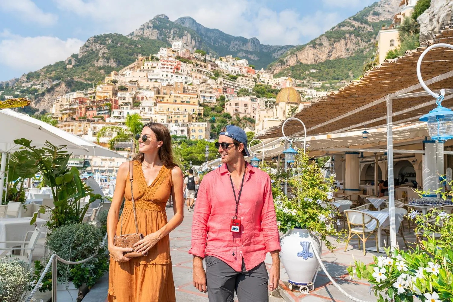 Two walkers on a terrace lined with plants and cafe chairs, Positano's hillside behind