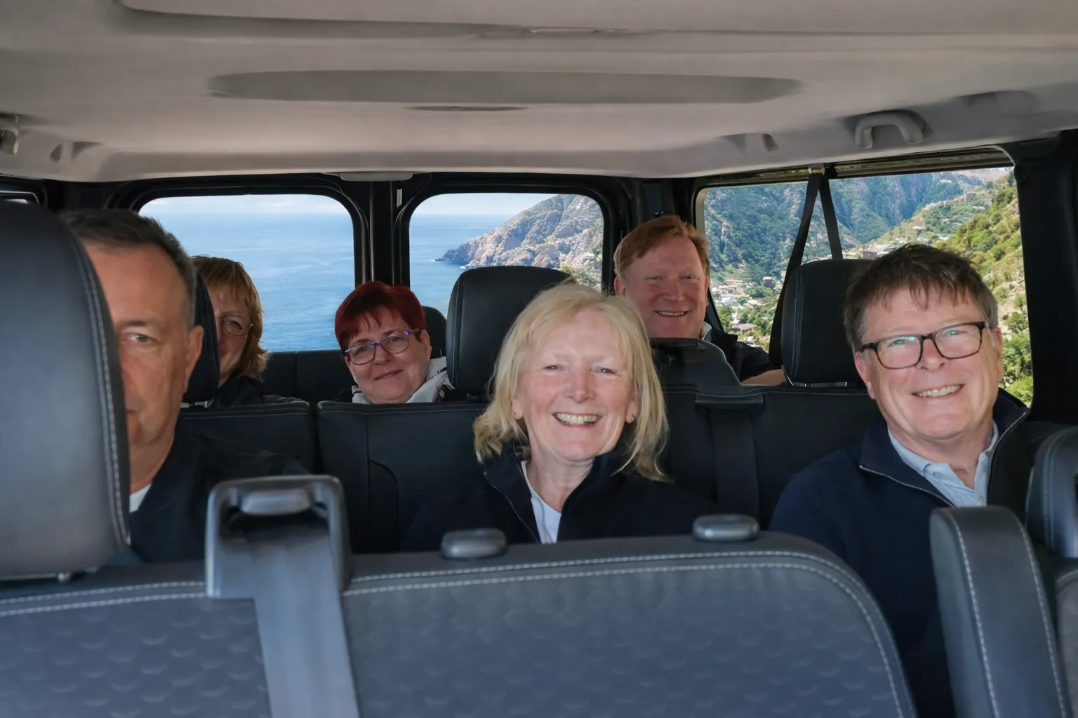 Passengers smiling inside a minibus, with sea and mountains visible through the windows