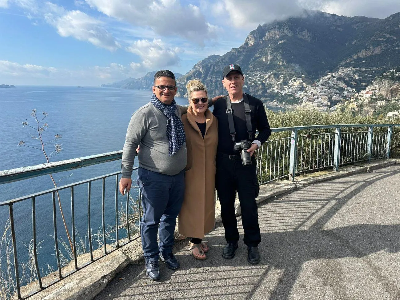 Three people posing at a railing on a coastal viewpoint with mountains behind