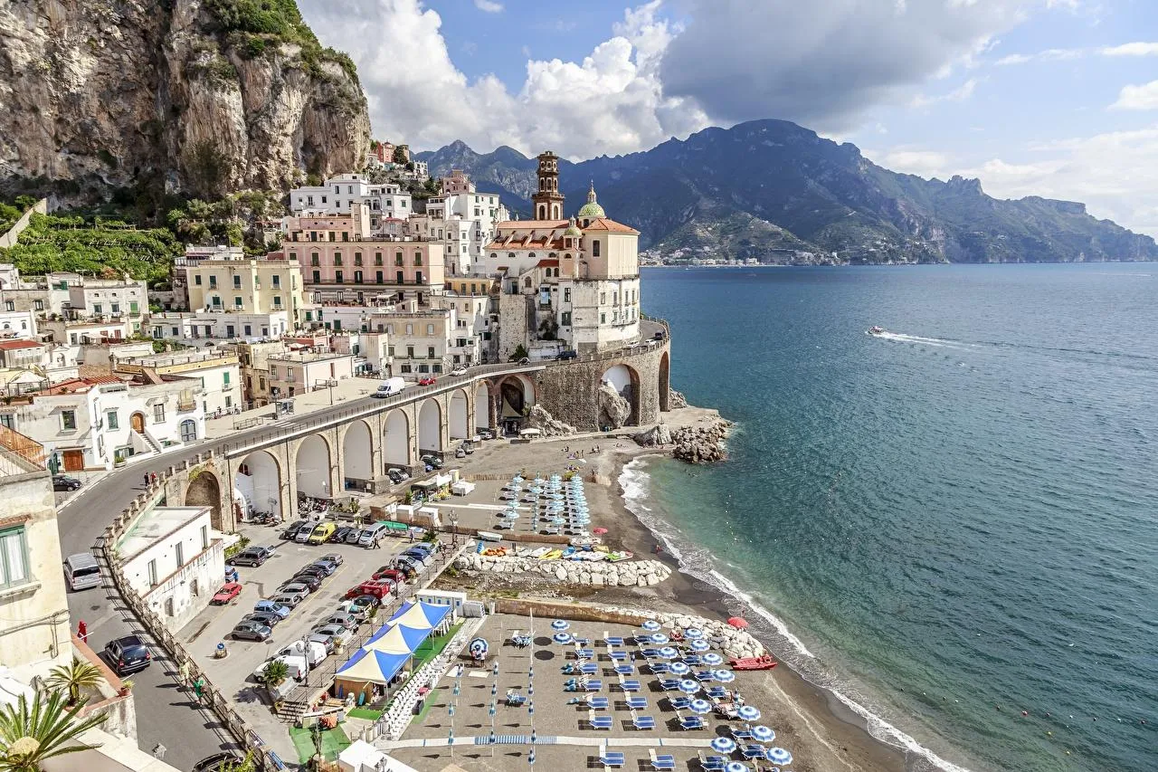 Atrani's houses and arched road above a beach with umbrellas and a calm sea