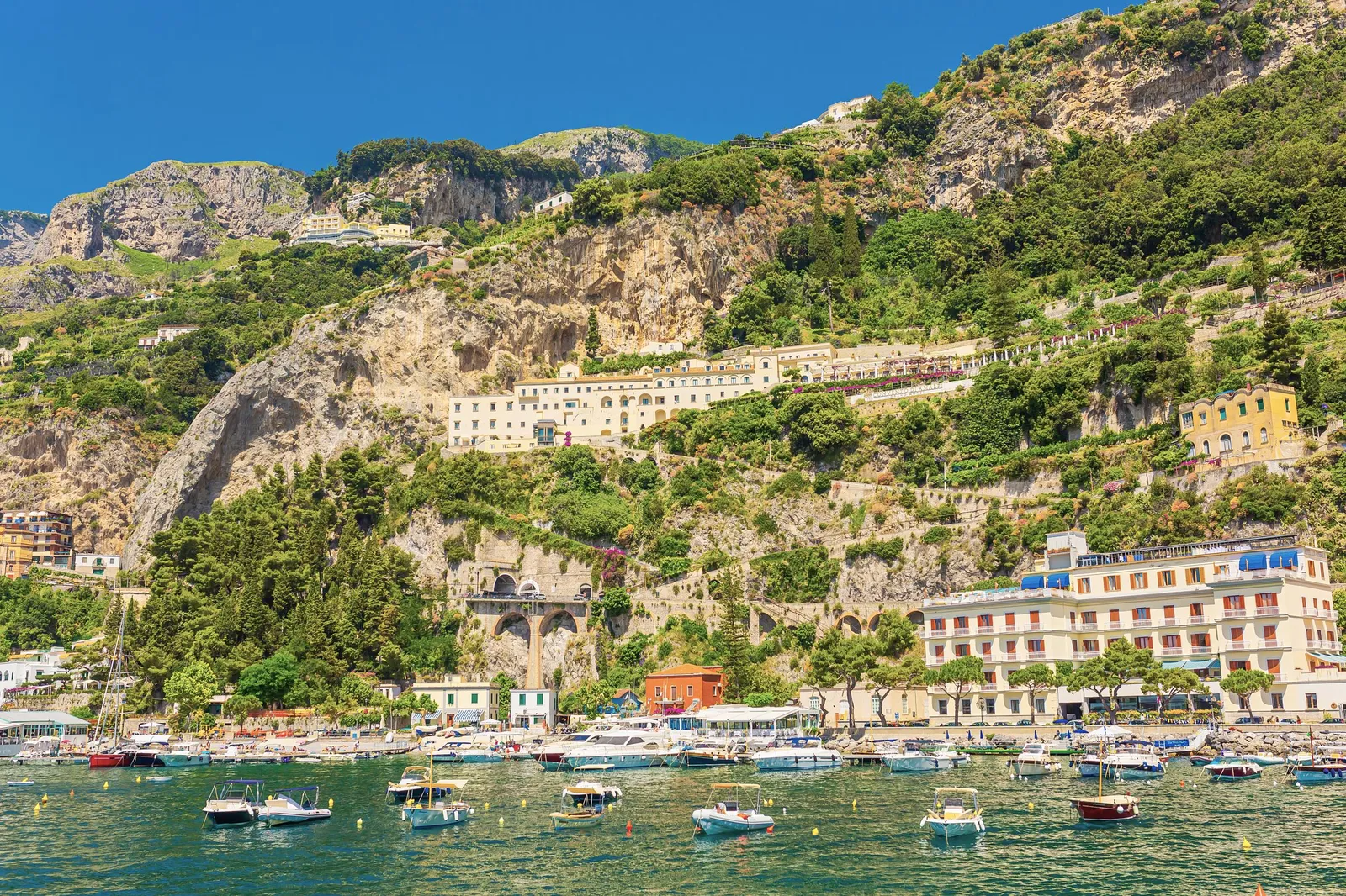 Boats moored in a harbour below hotels and terraced green cliffs