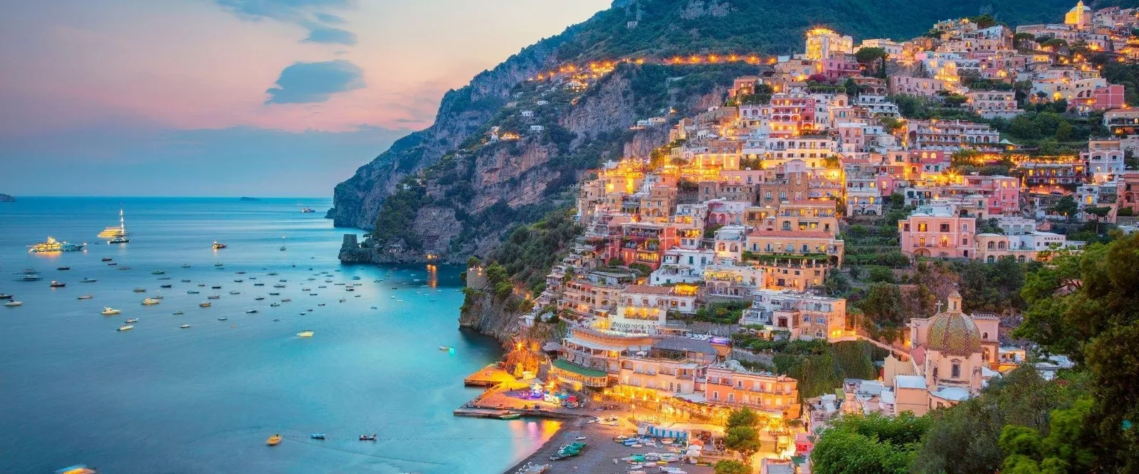 Positano's pastel houses stacked on the hillside at dusk, with boats moored in the bay