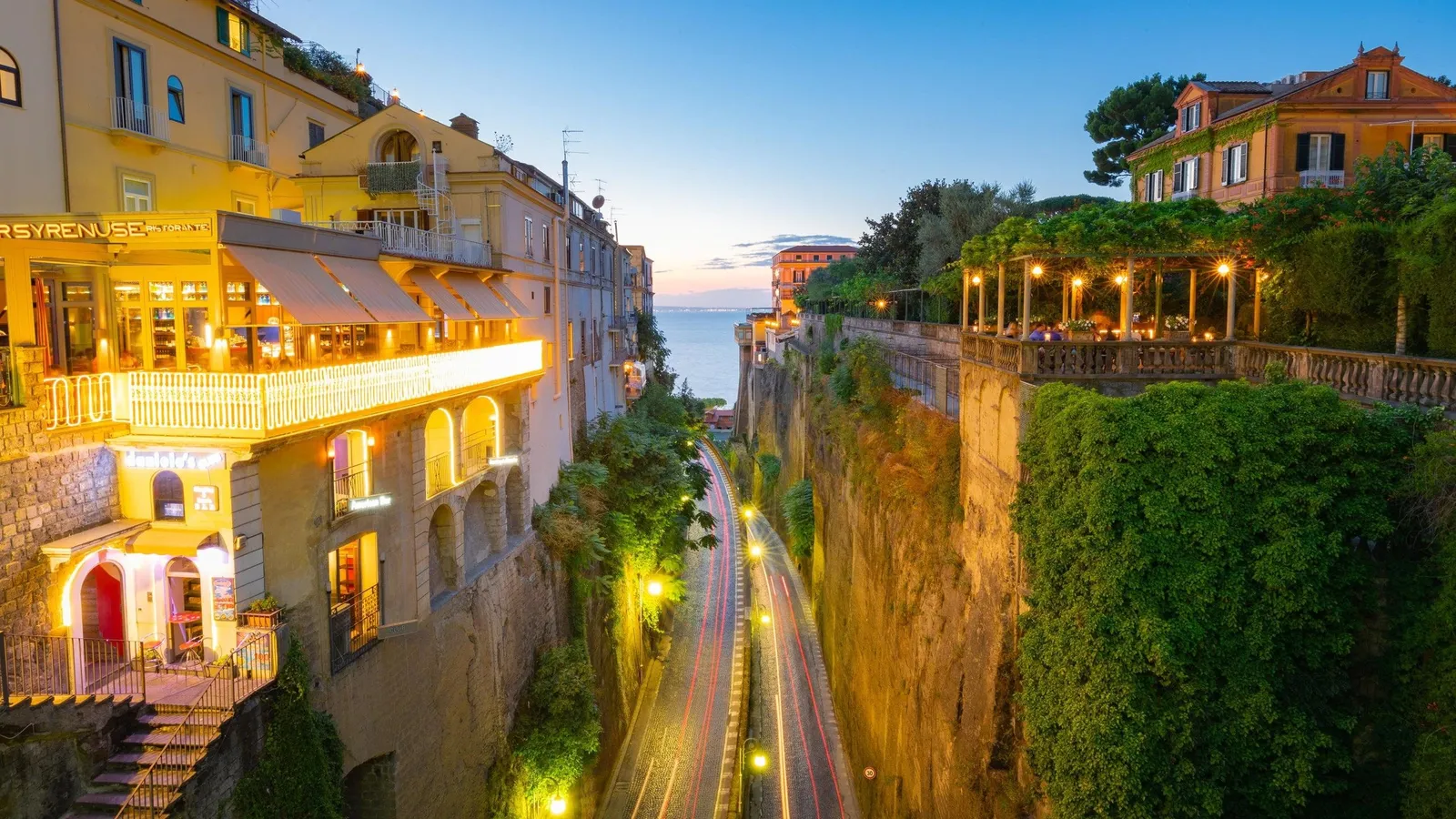 Lit restaurant terraces and a road in a cliffside gorge at dusk, sea beyond