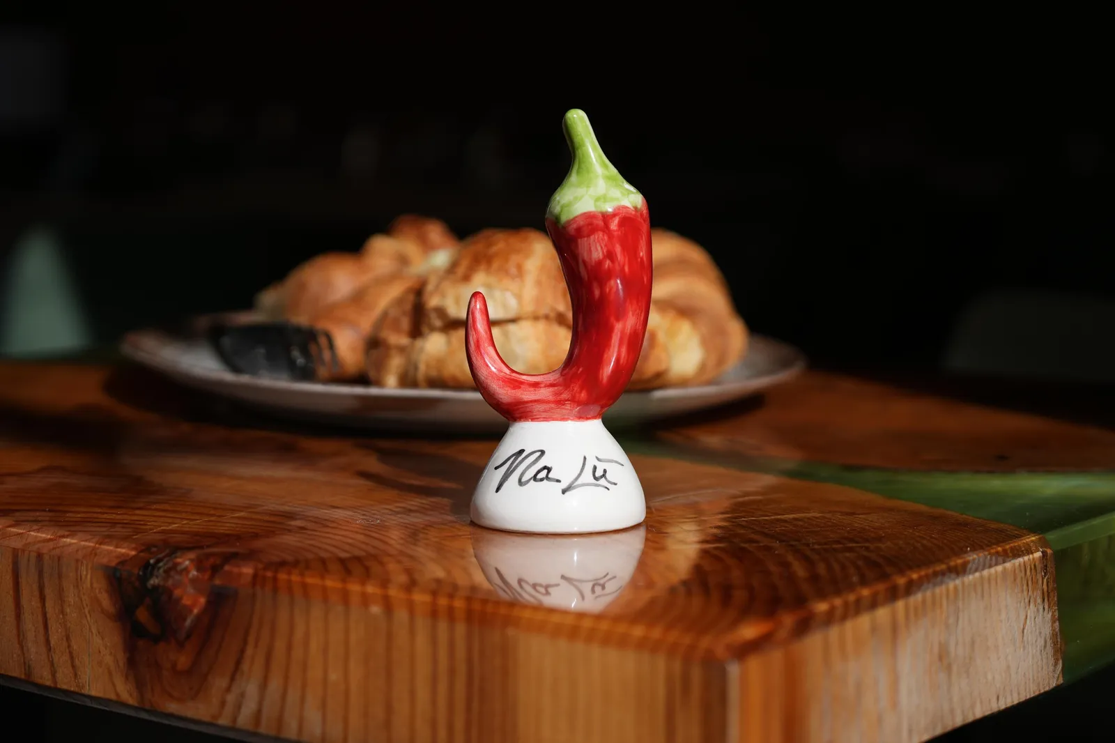 Ceramic red chili pepper figure on a wooden board beside a plate of croissants