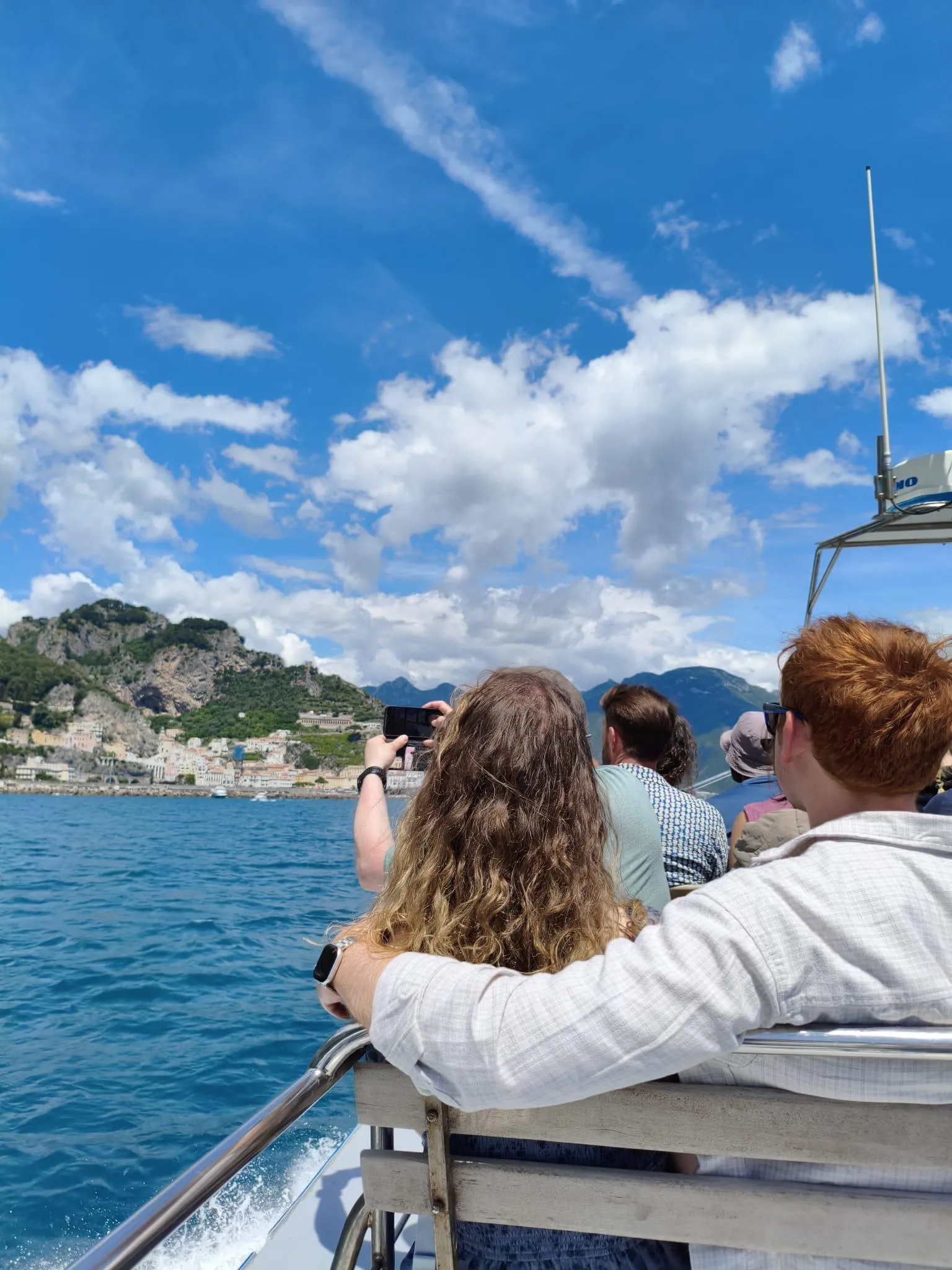 Two passengers on a boat deck with a green headland and blue sky ahead