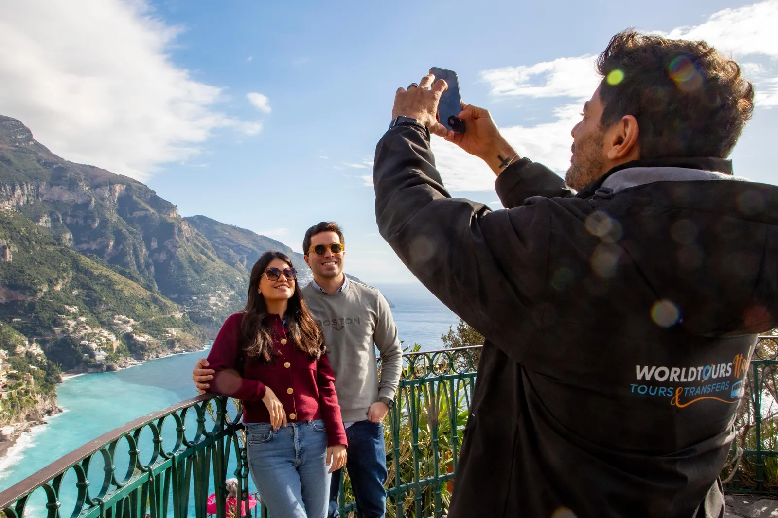 Man photographing a couple at a green railing above a turquoise coastline