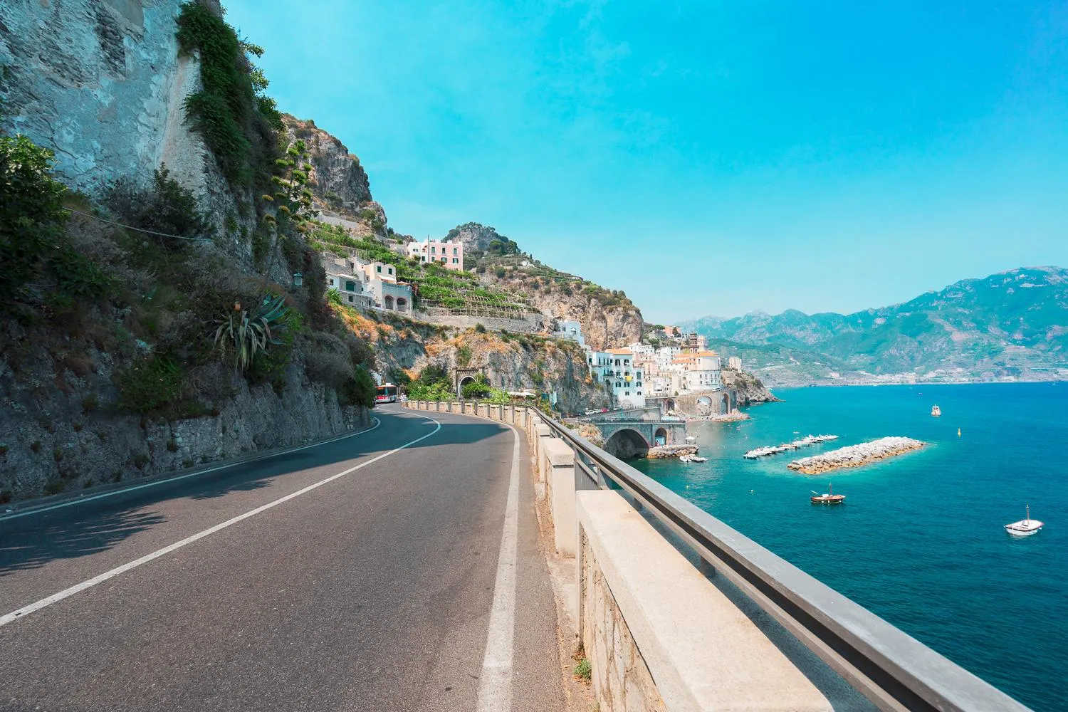 Coastal road with a guardrail curving above the sea toward a distant cliffside town