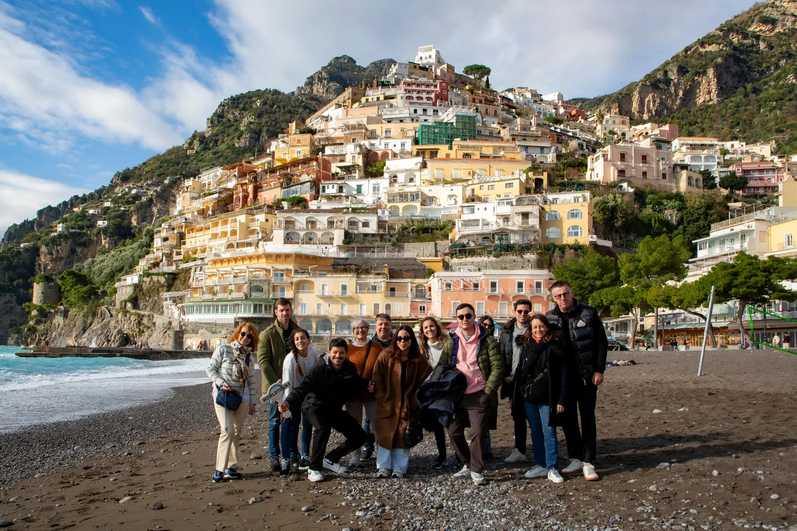 Group posing on a dark pebble beach below Positano's colourful houses